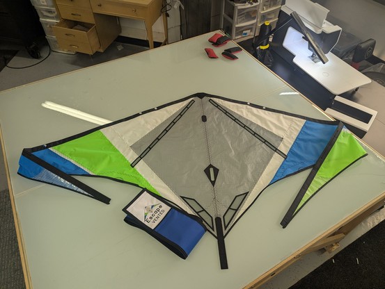 An unframed white, gray, green, and Blu vented Escape sport kite laying on a table alongside a custom bag for the kite.