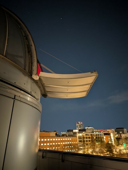 The exterior of a small, open, observatory against the night sky, with the Central Academic Building (CAB) visible in the background.