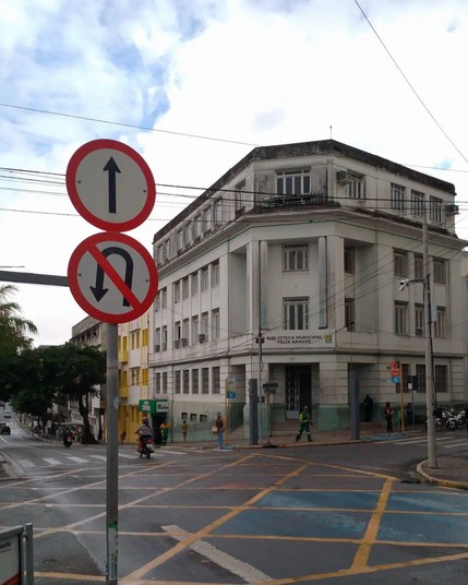 Um prédio branco antigo de quatro andares do outro lado de uma via de trânsito sem carros. Ele ocupa a esquina, portando, sua arquitetura faz uma curva. A frente há uma placa de trânsito informando "mão única" e "proibido retornar".