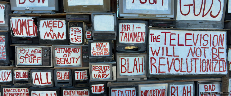 A collection of old televisions stacked together, each screen painted white with bold red text conveying critical messages about media, consumerism, and entertainment. Phrases include "THE TELEVISION WILL NOT BE REVOLUTIONIZED," "WOMEN NEED A MAN," "YOU NEED MORE STUFF," "STANDARD OF BEAUTY," "HOW LOVE WORKS," "RESULTS MAY VARY," and "IRRITAINMENT." The words highlight themes of societal expectations, advertising, and media influence. Some screens also display repeated words like "BLAH" and "BUY."