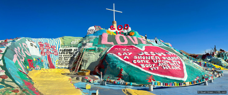 "An image of Salvation Mountain, a large, brightly colored art installation in the California Desert. The mountain is covered in vibrant, multicolored paint, with an array of religious and inspirational messages. At the top of the mountain, a white cross stands prominently. The front of the mountain displays a large message in red, stating "GOD IS LOVE," with other words such as "FAITH," "LOVE," "JOY," and "PEACE" painted across the surface. The paint forms patterns of flowers, stripes, and various shapes, creating a striking, whimsical visual effect. The background shows a clear, sunny sky, contrasting with the vivid colors of the mountain.