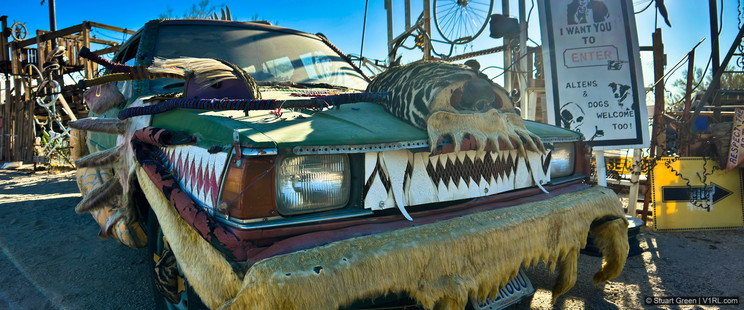 The image shows a creatively decorated car with various unusual and eye-catching elements. The car's front is adorned with a large animal skull, faux fur, and painted teeth, giving it a fierce and wild appearance. There are also colorful ropes and other decorations attached to the car. In the background, there's a sign that reads, "I WANT YOU TO ENTER. ALIENS & DOGS WELCOME TOO!" The setting appears to be an eclectic outdoor area with various artistic and mechanical structures.