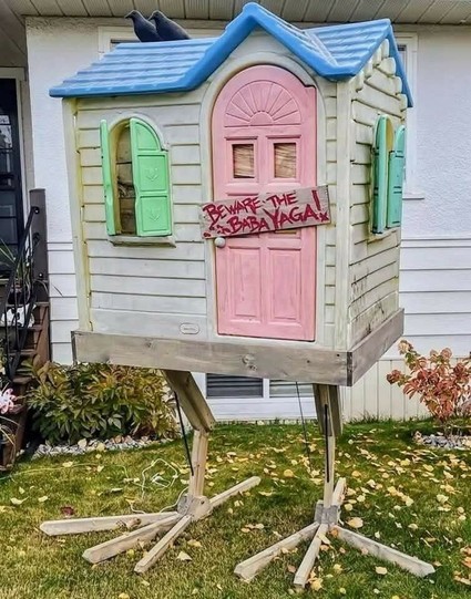 A plastic yard toy playhouse with  cute pastel accents has been altered: two ravens are on the roof and chicken feet (made of wood) hold the whole thing up. There's a wooden plaque on the door that says "Beware the Baba Yaga!" in all capitals & red ink