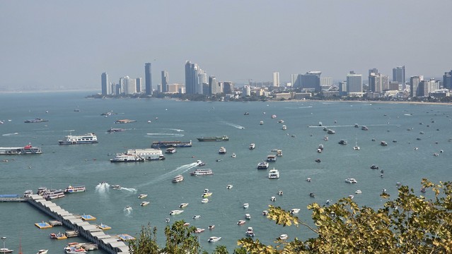 Blick von einer Anhöhe auf die Skyline von Pattaya (Hintergrund). 
Im Vordergrund ist ein Hafen mit vielen Booten und Yachten zusehen.