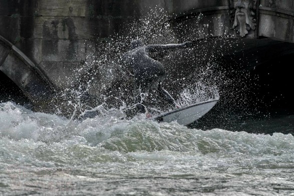 Farbfotografie im Format 3:2 Landscape.
Auf der stehenden Welle des Eisbaches in München versucht sich eine Surferin zu halten. Im Vordergrund ist die Welle, zu sehen, wie sie über die Sol Schwelle hochkommt. Im Hintergrund sind die zwei Kanalbögen zu erkennen, durch welche der Eisbach fließt.