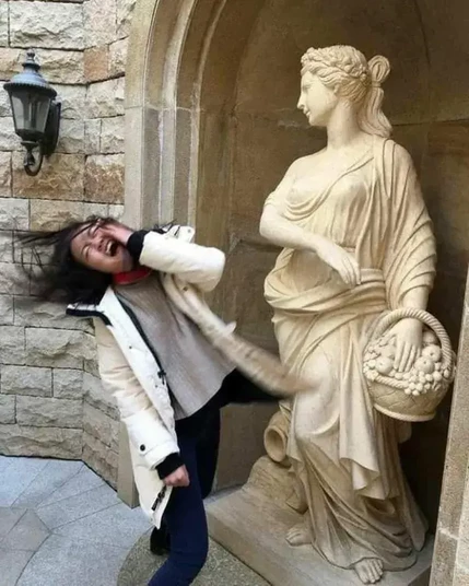 A perfectly timed photo of a woman appearing to be struck by a classical-style stone statue. The statue, depicting a robed woman holding a basket of fruits, is positioned in a niche of a stone wall, with an outstretched arm that aligns with the woman's head. Her shocked expression and windblown hair add to the illusion of impact, creating a humorous and dynamic visual moment. The setting suggests an elegant courtyard or historical architectural space.