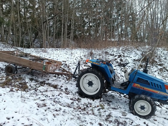 Blue tractor facing downhill on a snowy road that is sloping more towards the front wheels. The trailer is partially filled with snow covered logs. A forest is behind, on a level area.