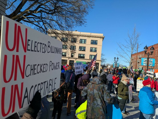 Protesters in Concord, New Hampshire. Lots of signs, lots of American flags, lots of chanting. The sign in the foreground says unelected billionaire, unchecked power, unacceptable.