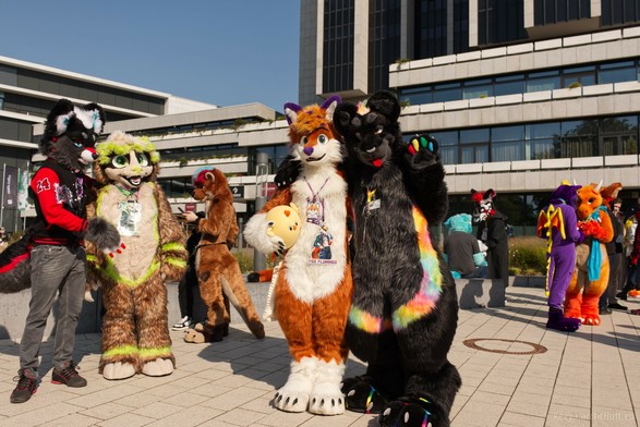 A photo from the courtyard of the Congress Center Hamburg during Euroference. Rodusk is standing next to Gustl and Fox is next to Kianga who has his paw over Fox's shoulder. Fox is holding a roumd chicken plushie