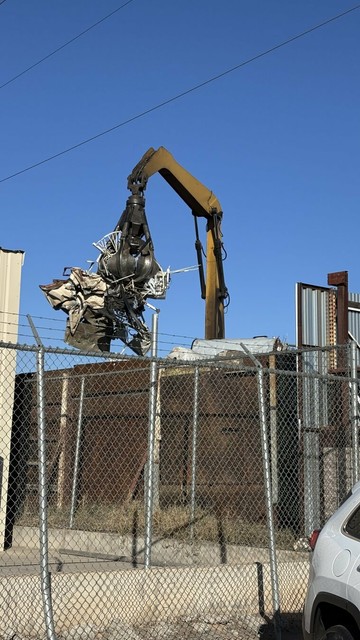 Heavy crane with claw lifting scrap metal, behind a metal fence, then a wooden fence, framed against a bright blue sky.