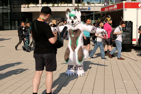A person in a white and grey fursuit with green highlights, not sure what species. They are standing with arms stretched out into a T shape in front of someone with an air blower.