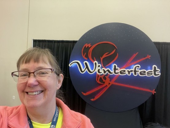 Woman with glasses happily next to big round Winterfest sign. Don’t let those skis fool you, it’s 60 degrees this year