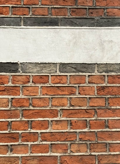 Rode bakstenen muur, met boven een brede horizontale witte betonnen strook, boven- en onderaand omrand door een rij van zwarte bakstenen