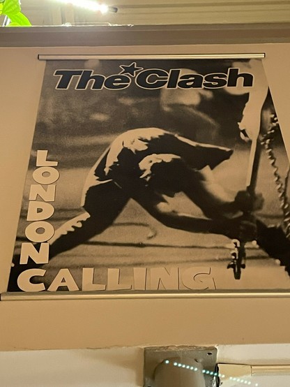Poster on my wall of a version of the Clash's London Calling  album cover but unlike the album, with the band name and title in black and white, but featuring the smae balck-and-whie cover photo pf Paul Simonon smashing his bass [guitar] on the stage.