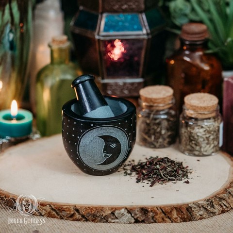 One black "Crescent Moon Soapstone Mortar and Pestle" product set stands on a raw wood slice. A pile of dried herbs sits next to it. The mortar is carved with a disc that contains a crescent moon with a face. The set is black, and the stone is lighter where it's been engraved. The inside of the mortar and the end of the pestle are the lighter color. Presented by Inked Goddess Creations.