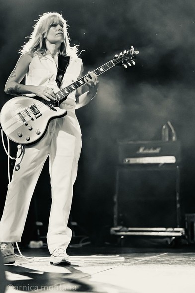 A guitarist in white clothing plays an electric guitar on stage, surrounded by a smoky atmosphere and illuminated by stage lighting. An amplifier can be seen in the background.