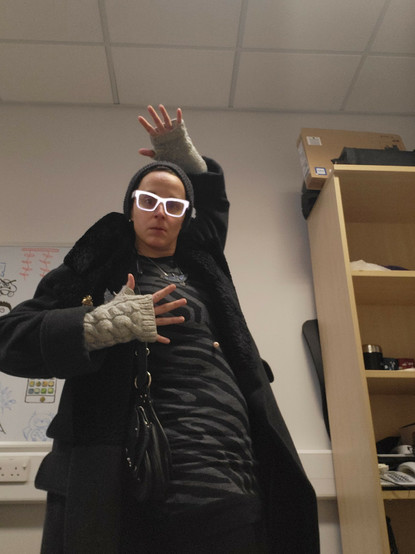 A fair skinned person wears a close fitting black knit cap, large reflective framed glasses, a voluminous black coat over a grey and black zebra print knit dress with a black leather holster style handbag and pale grey fingerless knit wrist warmers. They stand awkwardly in a bland office,  with their right hand raised behind their head and their left held at chest height.
