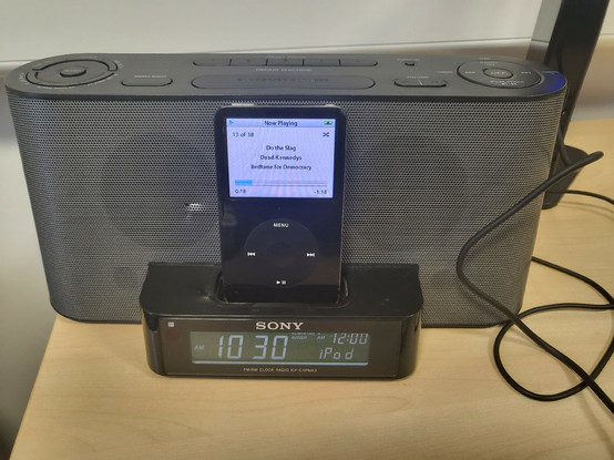 An old iPod, plugged into an iPod dock with speakers and a digital clock sits on a pale wood effect desk. Thermodynamics screen shows that it is currently playing "Do the slag" by the Dead Kennedys from their album Bedtime for Democracy.