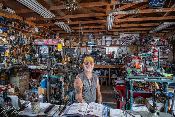 A mechanic sits at his table in a very filled work shop.