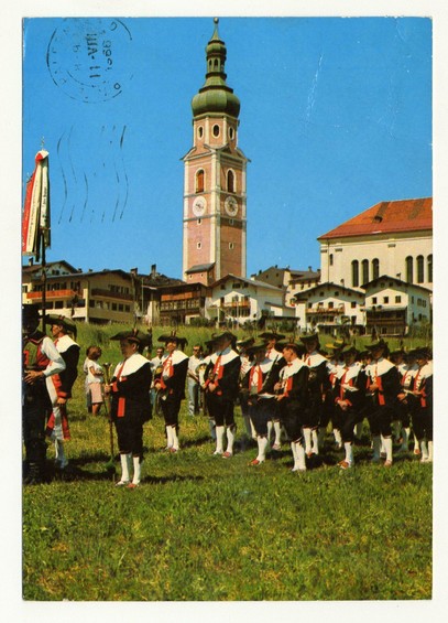 The image depicts a vibrant outdoor scene with groups of people wearing traditional Alsatian costumes, which includes black jackets adorned with red and white details. They are gathered in an open grassy area under a clear blue sky that serves as the backdrop for this setting. In the background stands a prominent clock tower painted in hues reminiscent of pinkish-red brickwork, topped by green accents on its dome-like structures.

Several individuals carry musical instruments such as flutes and drums, suggesting they might be part of a local band or orchestra celebrating an event like a festival or parade. The architecture behind them consists of buildings with red roofs that complement the overall picturesque setting typical for historical European villages in this region. Notably, there's also some sort of flag being held aloft on poles to add festivity and color contrast against the sky.

The postcard has stamps indicating it was mailed from Castelrotto Pr. Bolzano as noted at https://images.loener.nl/FerroCandilera/full/6730/6730eef889b10e7a26bbc58b.jpg, suggesting a historical context and possibly being part of an old collection or archive related to local customs in the Alto Adige region.
