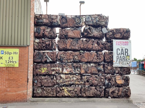Wall to a car park made up of blocks each of which are tightly crushed cars. A white and a yellow car park signs. The blocks are rusty coloured.