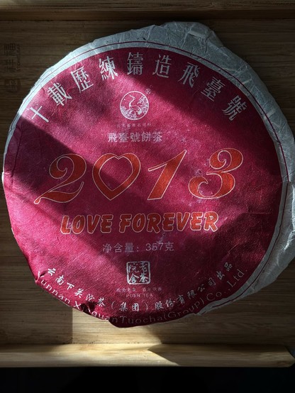 The wrapper of a cake of puerh tea. It has many Chinese characters around the outside, in the middle it has ‘2013’ in large print (the 0 is a heart) and ‘Love Forever’ underneath that. Above is the logo for the xiaguan tea company (a crane).