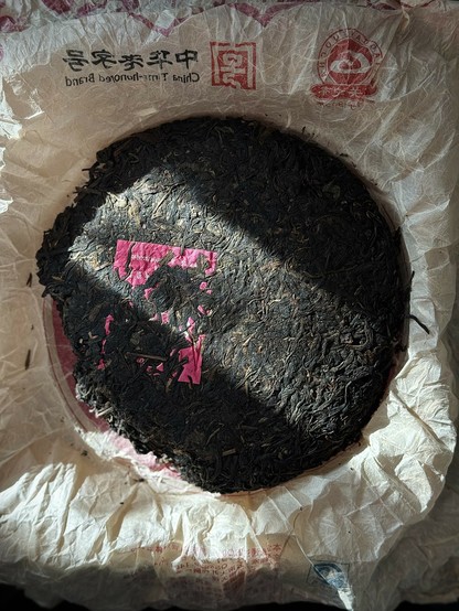 The face of a puerh tea cake with neifei half buried under pressed and dried tea leaves. The cake is sitting in its open wrapper with some Chinese characters at the top.
