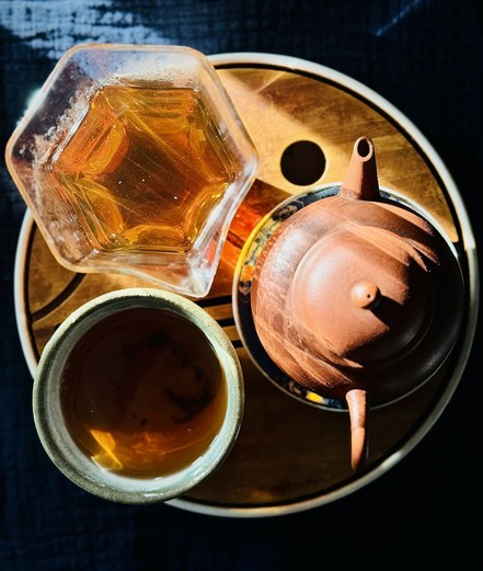 A yixing teapot, glass pitcher, and teacup arranged on a bamboo tray in the morning light. The pitcher and cup are filled with amber colored tea. The light streaming through the glass pitcher and onto the teapot.