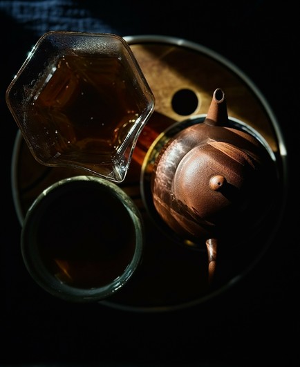 The same yixing teapot, glass pitcher, and teacup on a bamboo tray. The pitcher and cup are filled with amber colored tea. The scene is much darker with just the top of the teapot illuminated.