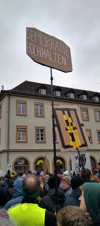 auf einem Teleskop-Stab ist ein Schild angebracht: Demokratie hochhalten; im Hintergrund ein mehrstöckiges Haus, unten einige Demonstranten