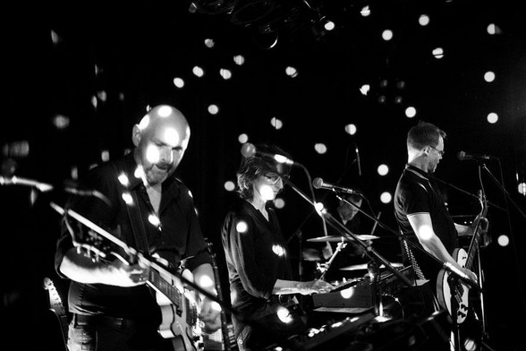 Das Schwarz-Weiß-Foto von Christian Schuller zeigt die Band VonLiebknecht während eines Live-Auftritts. Mehrere Musiker sind auf der Bühne mit ihren Instrumenten zu sehen, darunter eine Gitarre, ein Keyboard und ein Bass. Lichtreflexe in Form kreisförmiger Punkte sind über das Bild verteilt und erzeugen ein Muster auf den Musikern und ihren Instrumenten. Im Hintergrund sind Mikrofone, ein Schlagzeug und weitere Bühnenelemente erkennbar.