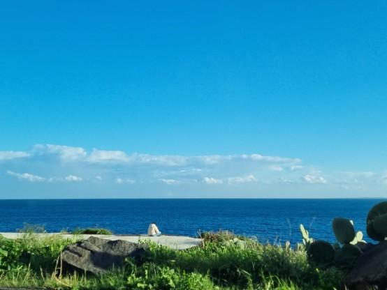 Cielo sereno dopo la pioggia, nuvole passeggere in lontananza, mare di un profondo blu, e di spalle una persona seduta ad ascoltare il mare o intenta a leggere