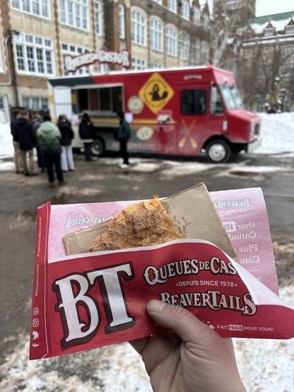 A person holds a Beavertail pastry, wrapped in branded paper, in front of a food truck. The truck is parked in a snowy area with a line of people waiting. The background features a historic building.