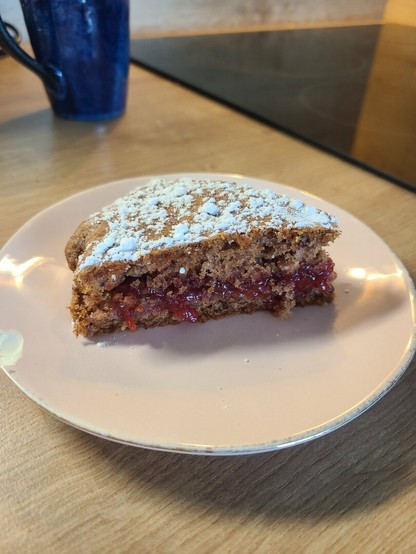 Photo d'une part de frucandie (gâteau végétal à la confiture) à la fraise, saupoudré de sucre glace. La part est dans une petite assiette rose un peu ébréchée sur le côté, et l'assiette est posée sur un plan de travail aspect bois clair. On distingue au fond un mug bleu et une partie de la plaque de cuisson.