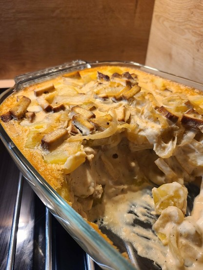 Photo en assez gros plan d'un plat à gratin contenant une tartiflette végane entamée. On voit des bouts de lardon de tofu fumé et des lamelles d'oignon mélangés aux pommes de terre et à la crème fromagère.