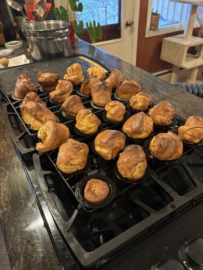 A collection of golden-brown popovers arranged on a baking rack on a stove, with a pot and kitchen utensils visible in the background.