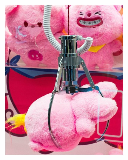 A pink stuffy held by a claw machine claw with laughing stuffies in the background.