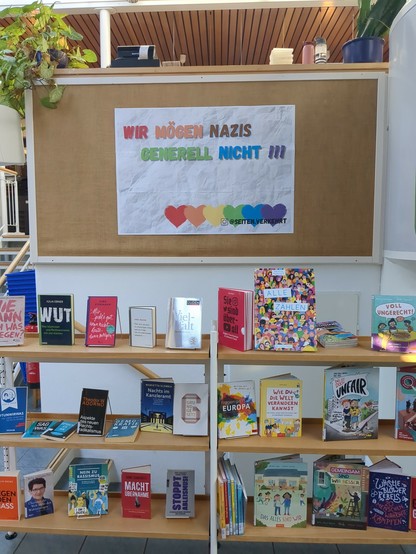 Ein Foto aus der Stadtbibliothek Rotenburg (Wümme)
In der oberen Hälfte des Bildes ist ein großes Plakat mit dem Text "wir mögen Nazis generell nicht!!!"
Jedes Wort ist dabei in einer andern Farbe. Unter dem Text sind Herzen in verschiedenen Farben.
In der unteren Hälfte des Bildes ist ein Regal mit Büchern zu sehen. Die Bücher haben Titel wie "Vielfalt", "Wie du die Welt verändern kannst" und "Nein zu Rassismus"