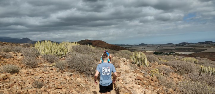 A column of runners on a dusty rocky trail, surrounded by dry shrubs and cacti, under a partly cloudy sky.