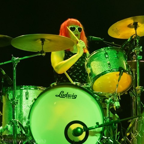 A drummer with bright red hair and white sunglasses performs on stage, playing a silver Ludwig drum set. The scene is illuminated with green lighting, highlighting the energetic atmosphere.