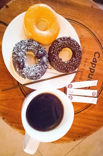 Photo shows three donuts on a plate with two types of dark chocolate coating, and one with sugar glaze. They are flanked of a white cup filled with black coffee. Small packets of sugar is on the plate of the coffee cup. The two plates are placed on a wooden table with some old coffee related inscriptions.