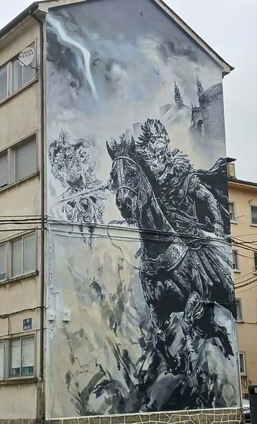 Mural/Grafitti cubriendo fachada del lateral de un edificio. Legionario romano a caballo, enzarzado en posible combate con otro jinete al fondo, bajo un cielo nublado.