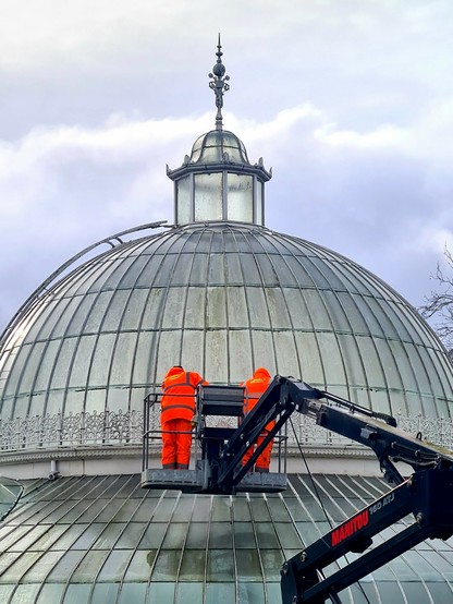 Two men in bright orange pressure-washing the windows of a Victorian glasshouse from a cherry-picker.