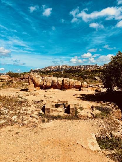 An ancient Greek statue lying down with the head between elbows on a boiling hot day starring at the town of Agrigento beneath a blue sky