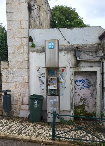 Um telefone público à frente de uma parede degradada, num passeio de calçada preta e branca. Ao lado do telefone encontra-se um caixote do lixo.