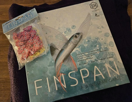 Die Box des Spiels auf einer Decke angerichtet. Dazu geliefert wurden die Deluxe-Token: Fische, Fischschulen und Fischeier.