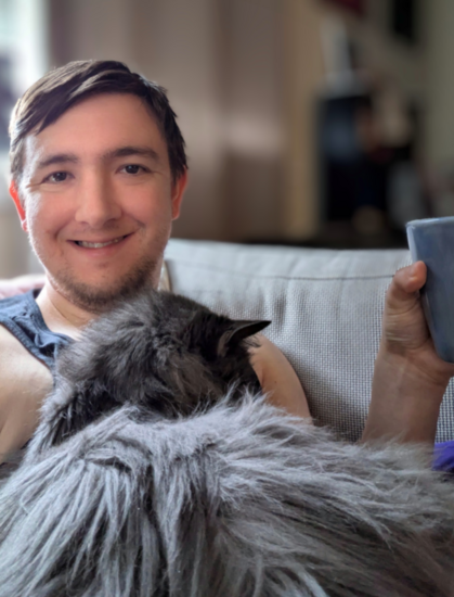 Man smiling wearing a black tanktop and holding a coffee mug sitting in an armchair. Oscar the cat is a fluffy gray/black nebelung purring facing away from the POV sleeping peacefully on the man's body.