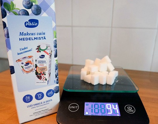 Valion mustikkakeitto purkki, jonka kyljessä teksti "Makeus vain HEDELMISTÄ"  

tekstin alla pienempi kuva, jota tarkemmin katsomalla näkee, että siinä on kuva jostain luonnonjugurttipurkistä, jolla ei ole mitään tekemistä tämän purkin mustikkakeiton kanssa.

Vieressä keittiövaaka, jolle on kasattu tuoteselosteen mukaiset 94 grammaa sokeria, eli 25 sokeripalaa.
