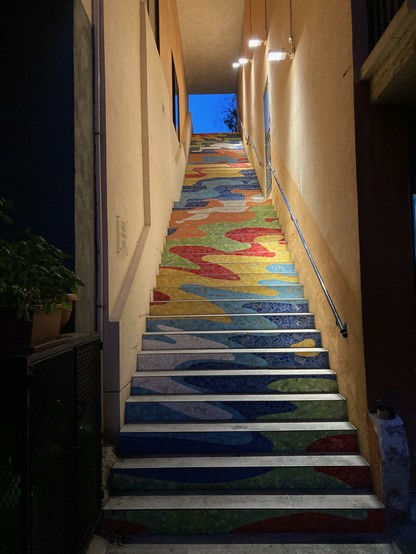 Eine bunte Treppe. Graue Trittflächen, aber die Vorderseiten mit bunten Kachelbruchstücken. Aus den Bruchstücken ergibt sich ein Wellenmuster. Blick geht aufwärts, in den dunkelblauen Abendhimmel.