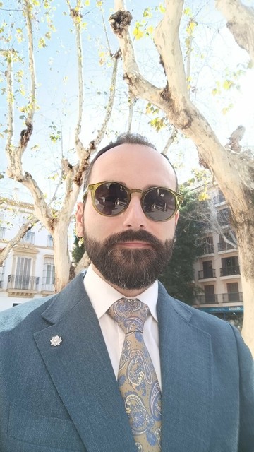 Selfie vistiendo un traje verde azulado con una camisa blanca y una corbata de diseño pashley en tonos caqui y azul. Llevo gafas de sol redondas y un pin de brillantes en la solapa del saco. El fondo muestra árboles altos con hojas dispersas y edificios residenciales.